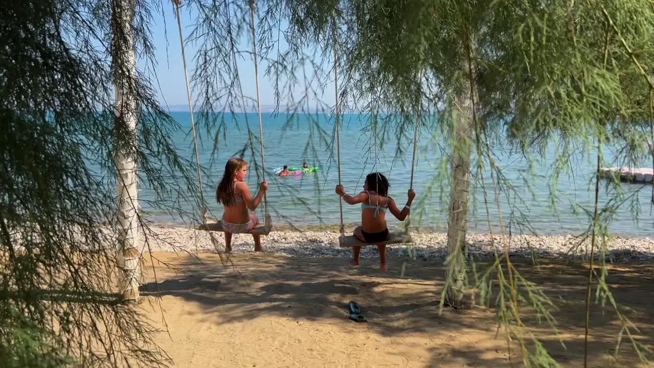 Back view of two little girls on vacation having fun swinging on seafront rope swing with tree fronds in foreground