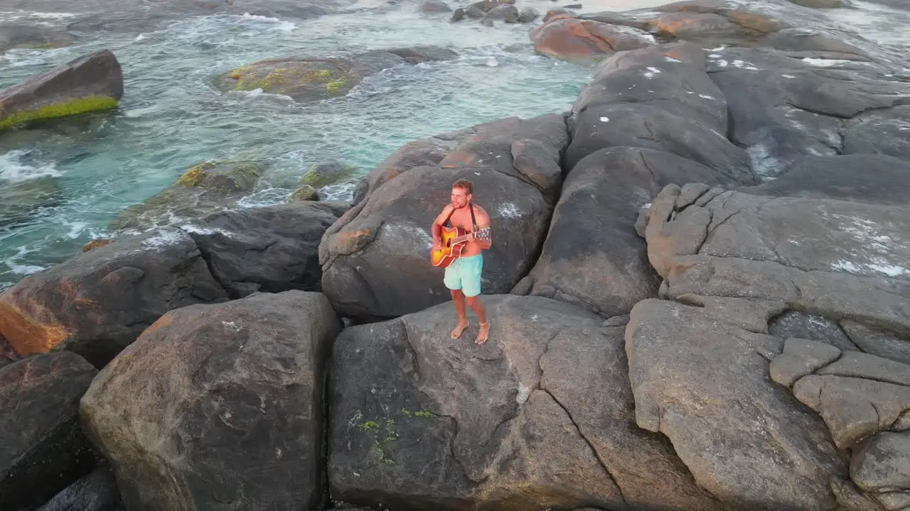 Aerial top down shot of young man in bathers standing on rock and playing guitar in front of ocean and beautiful sunset Australia