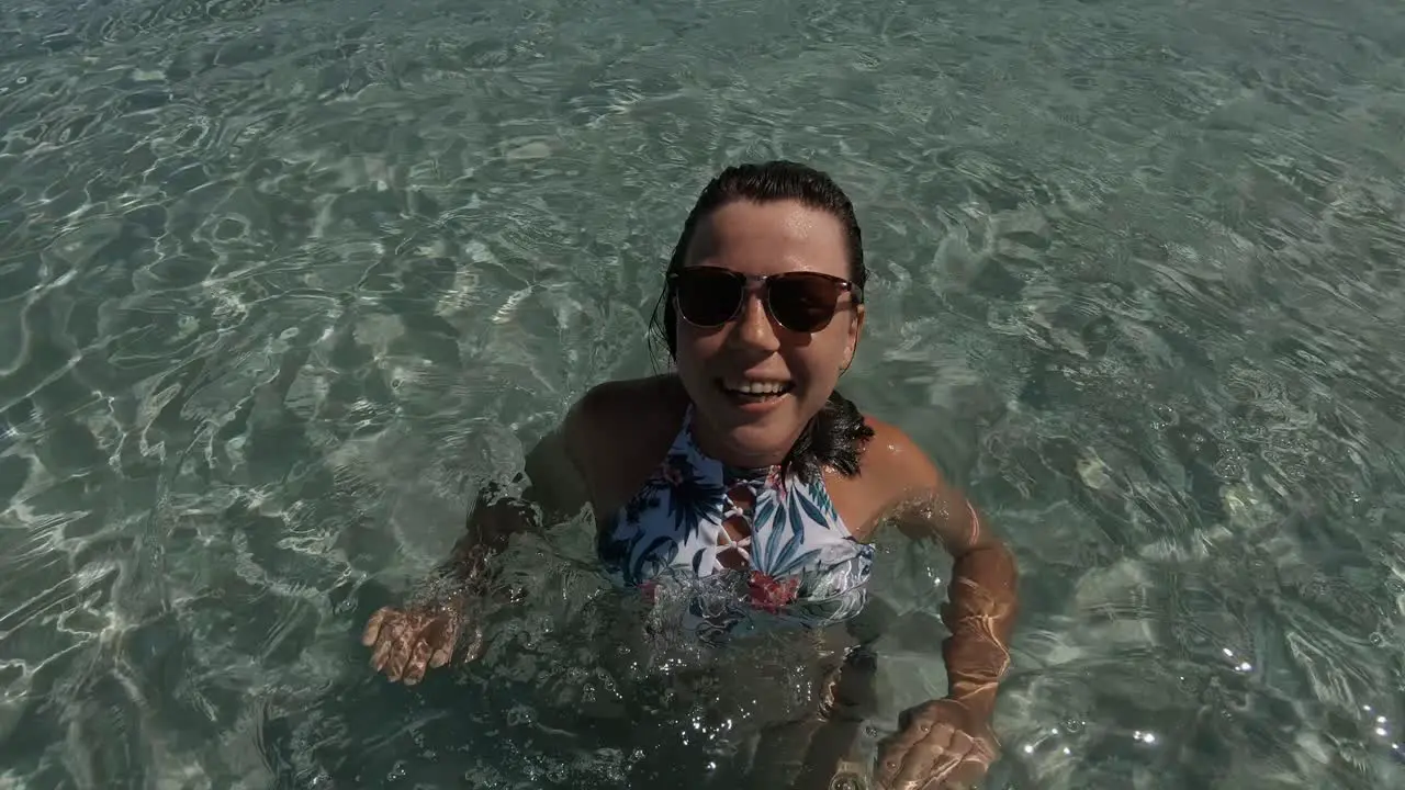 A pretty young girl in tropical swimwear and sunglasses fixing her long brown hair while smiling towards the camera