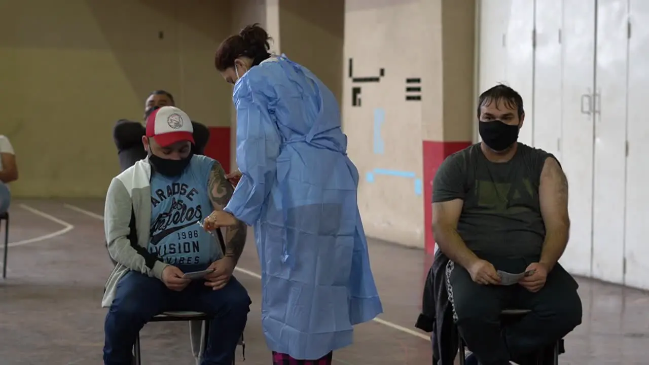 Medium close shot of a nurse applying a shot of COVD-19 vaccine to a tattoed latino man inside a makeshift vaccination center