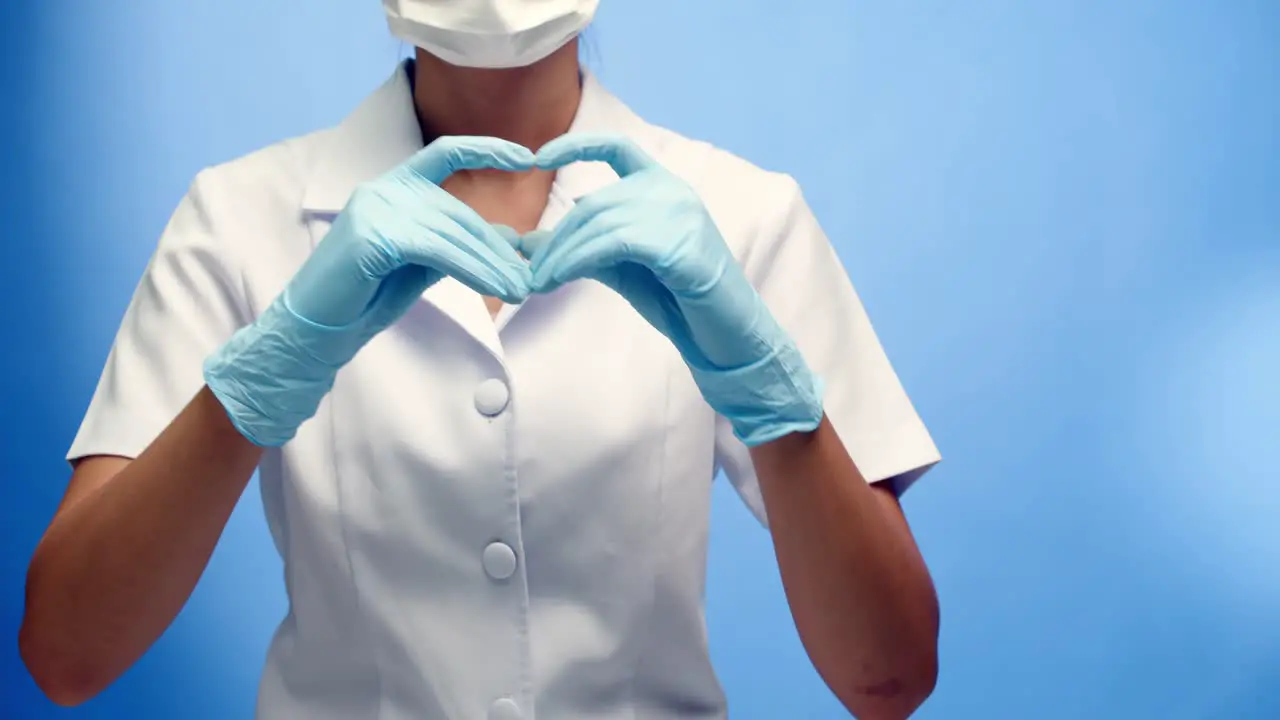 A nurse wearing a face mask and white uniform makes a heart sign with her hands blue background with copy space