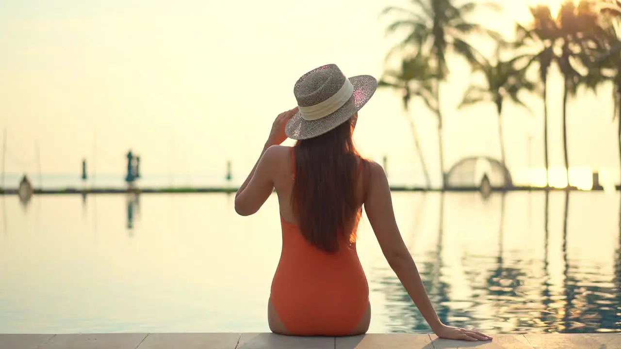 Back view of Fit female model wearing an orange monokini swimwear suit and hat sits on the edge of the swimming pool in Turkey at sunset