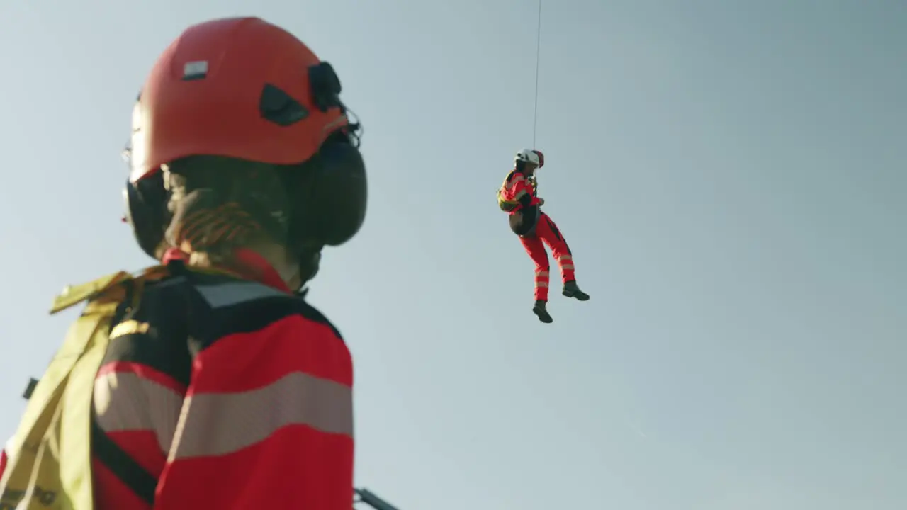 Rescuer hanging in the air from Helicopter hook and other one standing in foreground sunny slowmotion