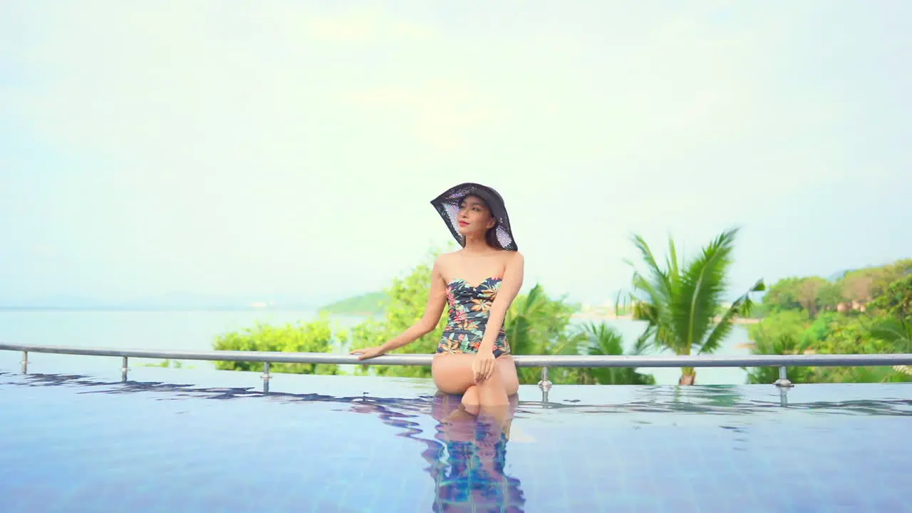 Asian woman in swimsuit sitting on pool edge and looking around with satisfaction