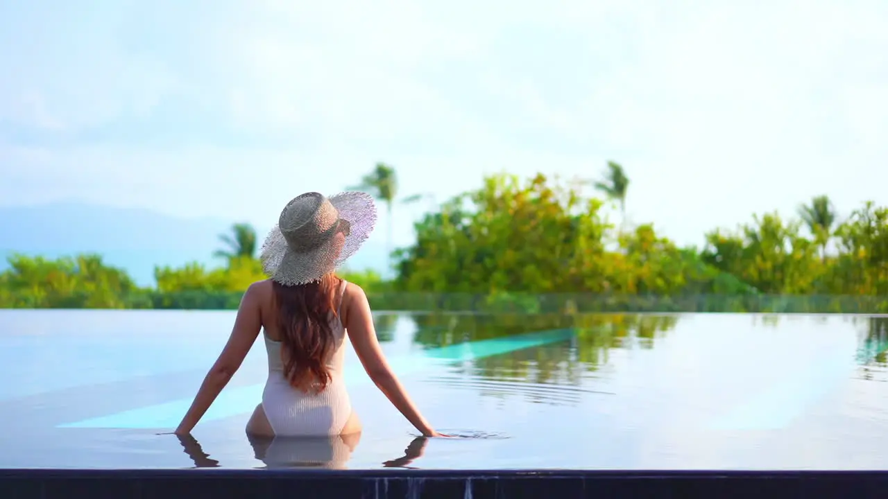 Back view of woman with hat sitting on infinity pool edge looking at panorama