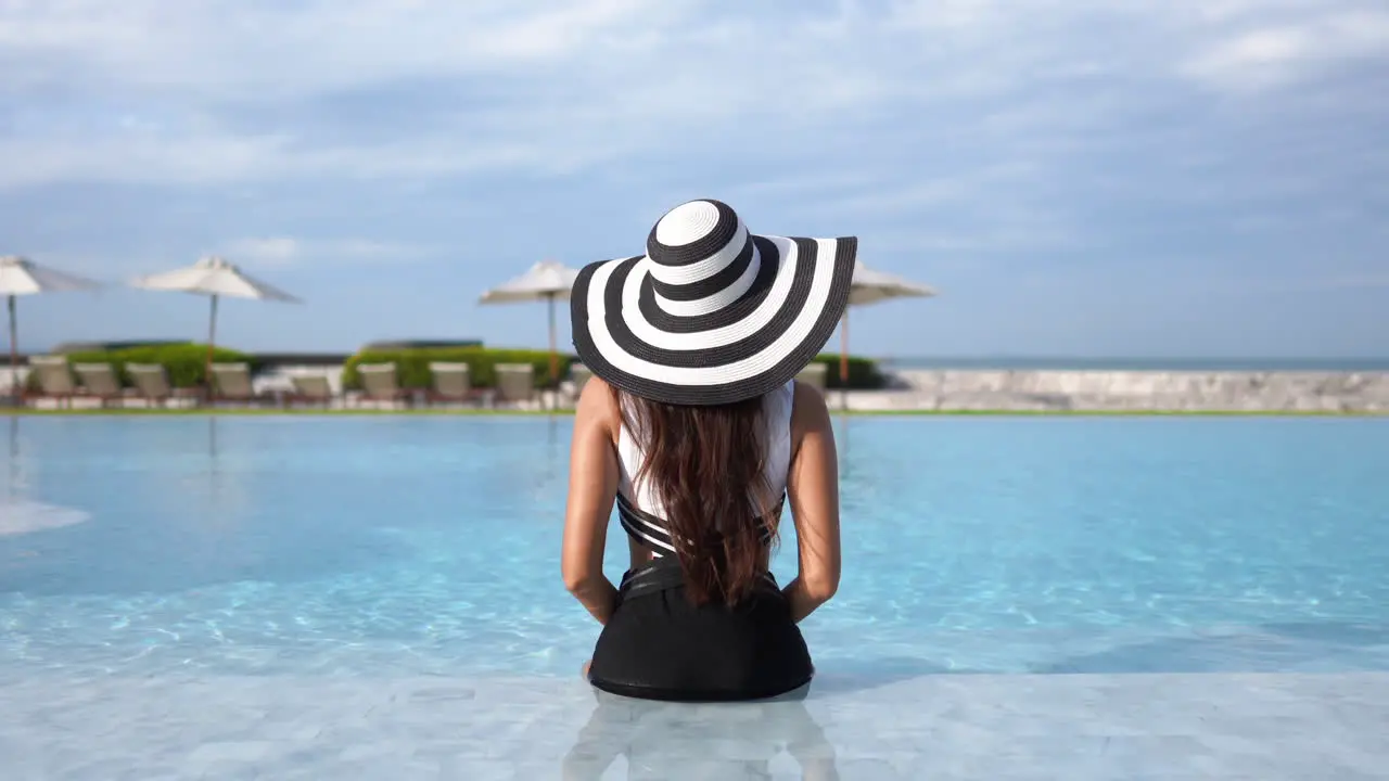 Static Shot of Back of Stylish Female With Black and White Hat and Swimsuit Sitting on Pool of Luxury Hotel Copy Space