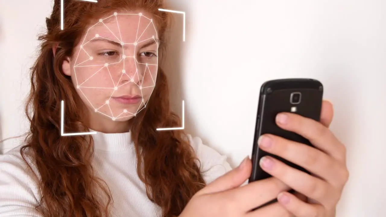 Close up of a young woman with red hair with a white shirt on a white background while holding a phone in front of her face with facial recognition high tech animation with a glow effect on her face