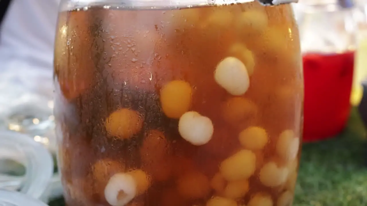 Food vendor stirring longan juice with fresh longan flesh in big glass jar at fruit juice stall at walking street