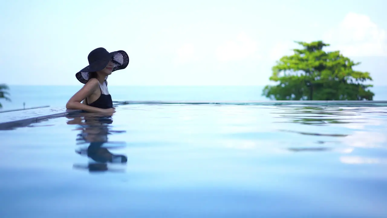 Elegant lady wearing a black sun hat leaning peacefully on the edge of a relaxing infinity pool as crystal clear water ripples with her reflection