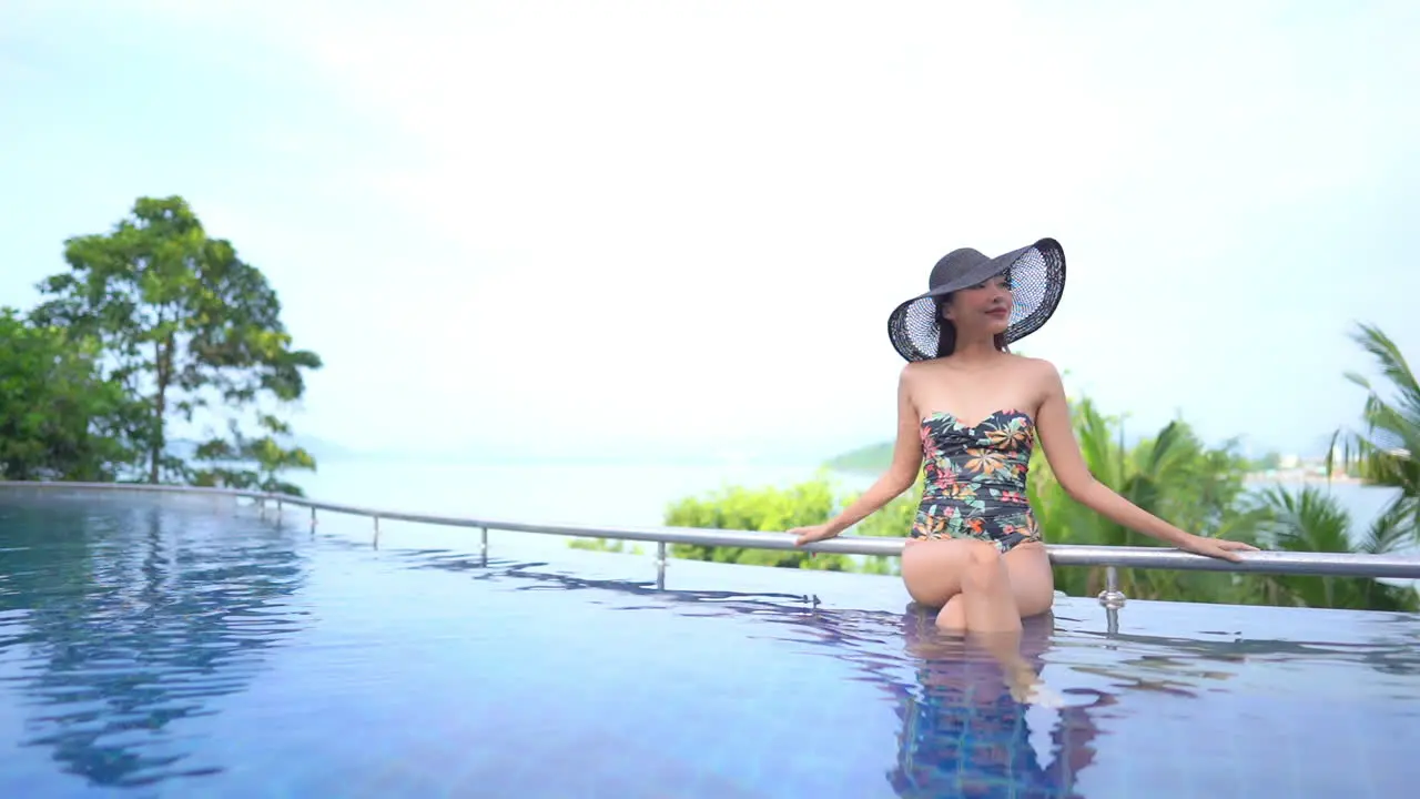 An attractive woman in a 40s Joan Crawford style sun hat sits along the edge of an infinity-edge pool with the ocean and a modern city skyline in the background