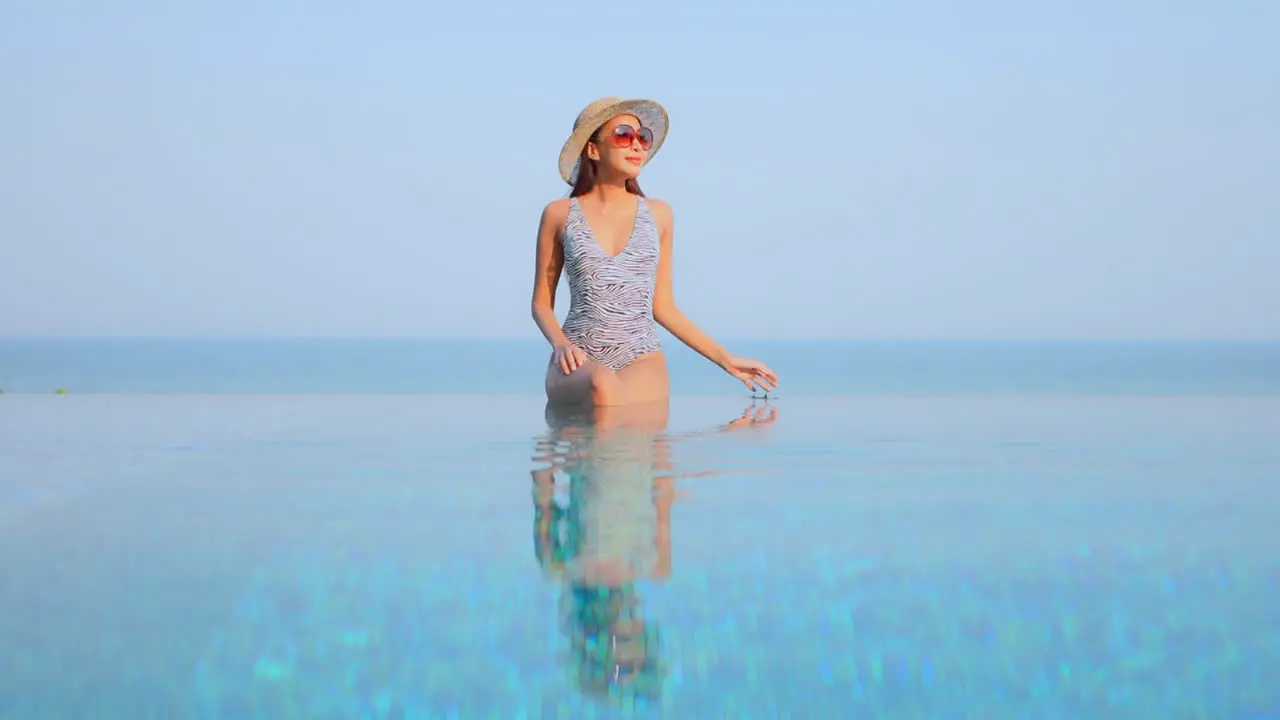 Pretty woman in swimwear sitting on infinity pool border touching her sunhat and raising hands up with a stunning view of tropical sea horizon in Phuket Thailand static copy space