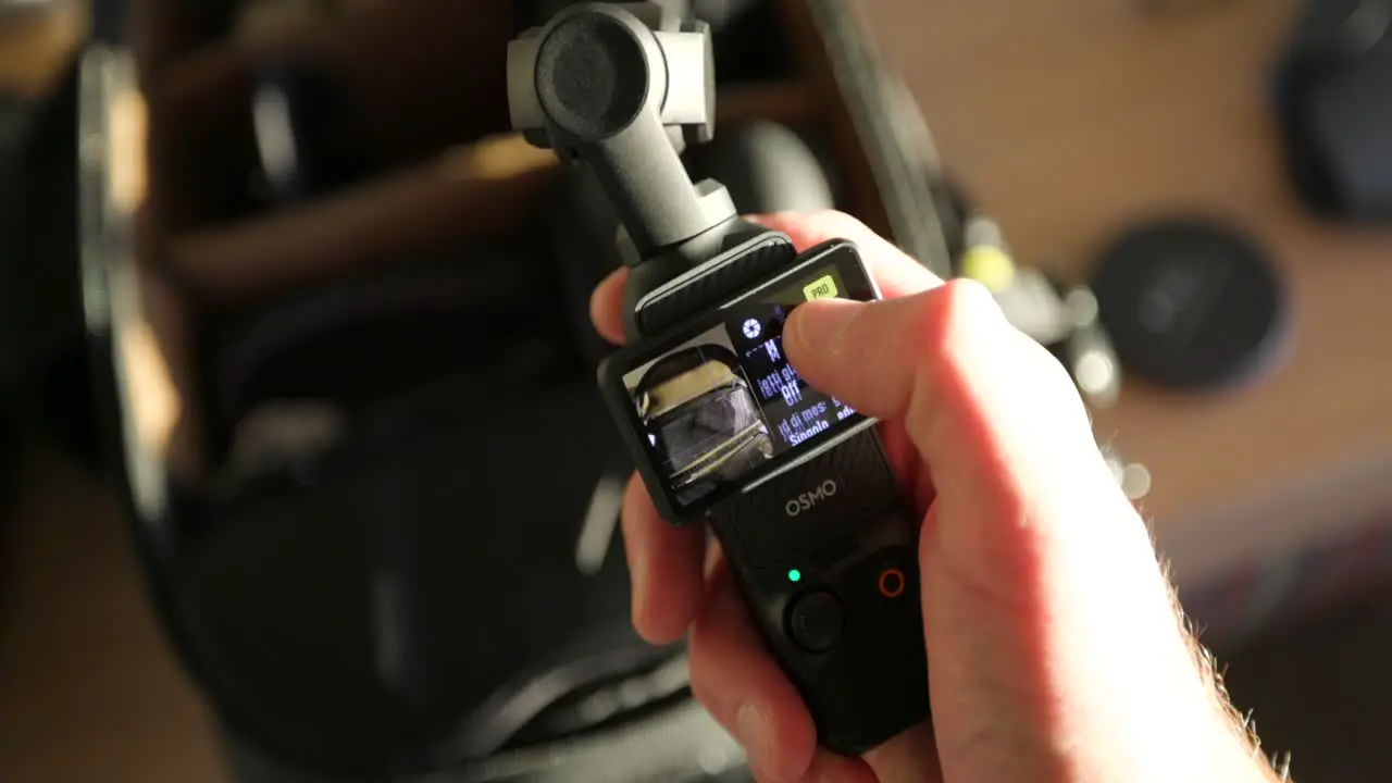  DJI Osmo Pocket 3 Held In Hand And Screen Interface Being Used With Bokeh Background Of Camera Bag On Table Indoors