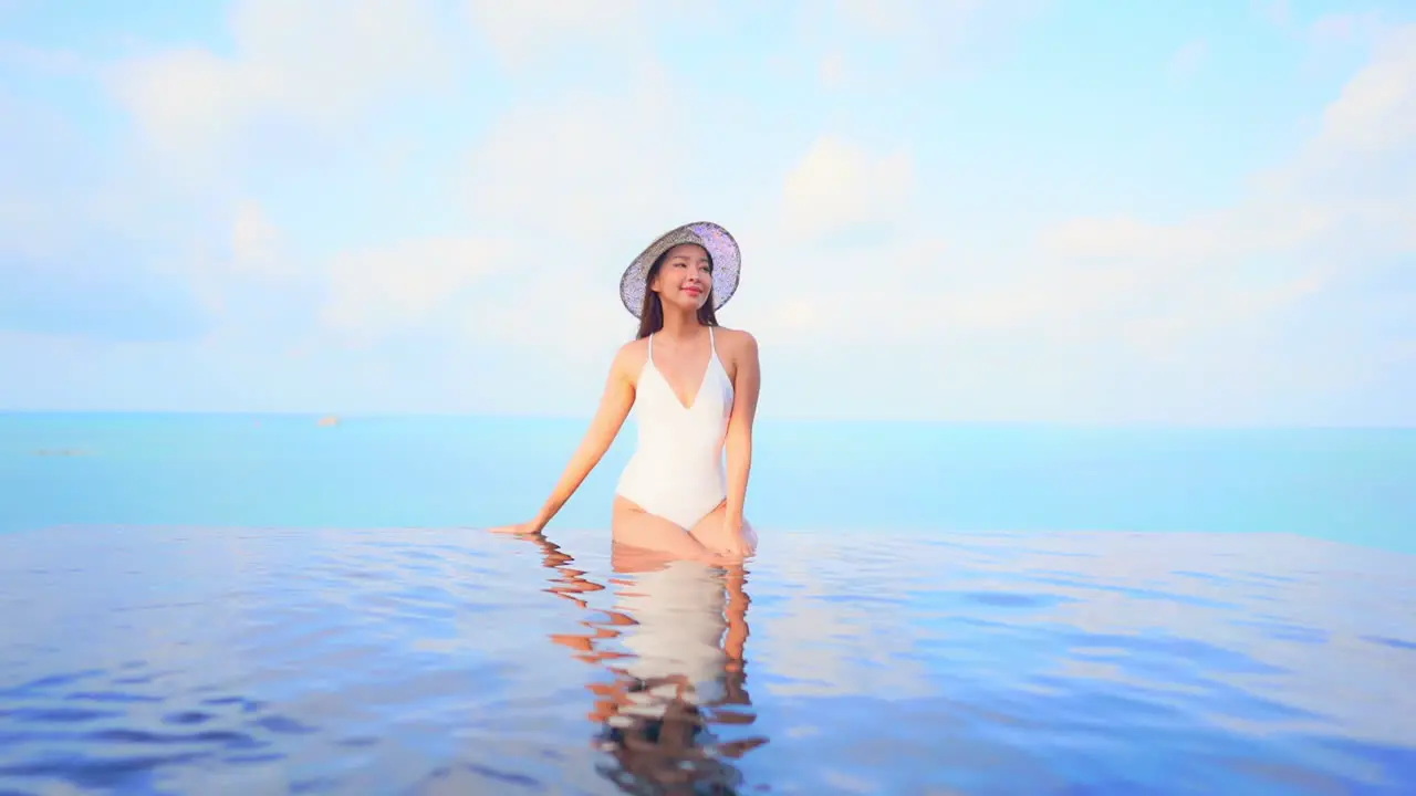 Stunning woman in a white bathing suit sits on the edge of an infinity-edge pool ocean in the background