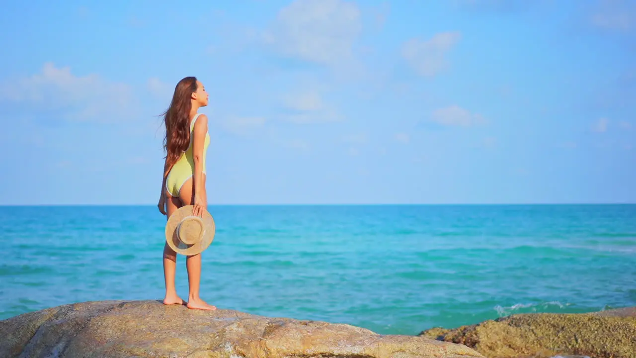 Standing on a large rock a pretty woman in a bathing suit and sun hat in hand looks over the ocean waves