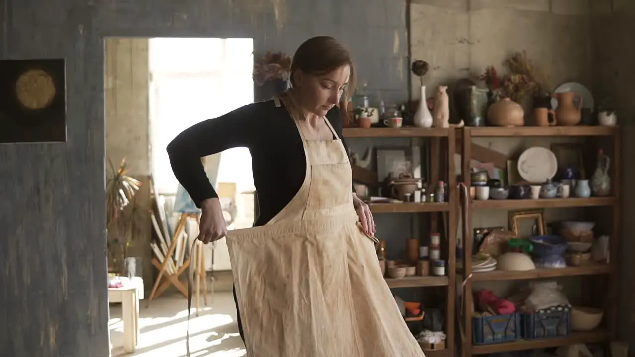 Slow Motion Footage Of Young Woman In Casual Clothes Standing In Pottery Workshop With Ceramic Wares And Supplies On Shelves And Table With Tools In Middle And Putting On Beige Apron