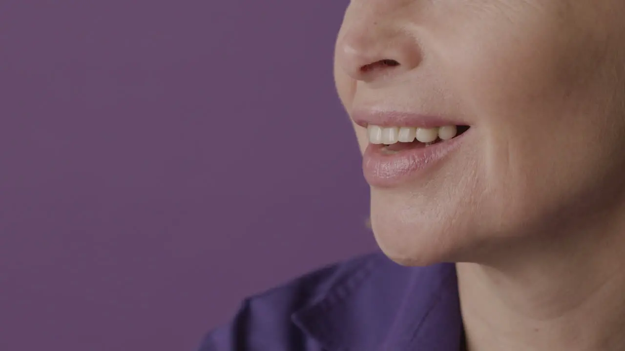 Close Up View Of Half Face Of Blonde Mature Woman With Purple Shirt Smiling On Purple Background