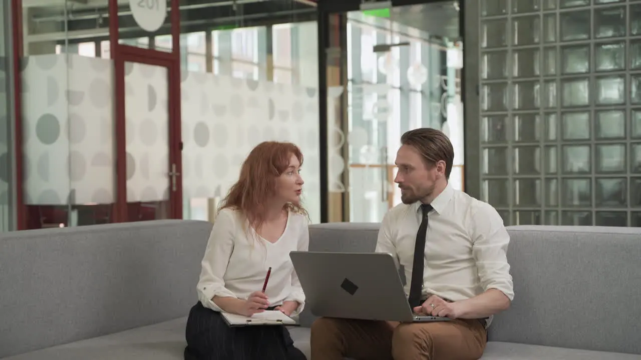 A Red Haired Businesswoman Has A Relaxed Meeting With A Young Man With A Laptop On The Couch In The Common Areas Of The Office Building 1