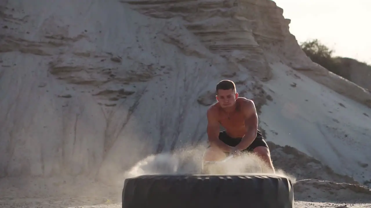 Muscle athlete strongman man hits a hammer on a huge wheel in the sandy mountains in slow motion at sunset The dust from the wheels rises