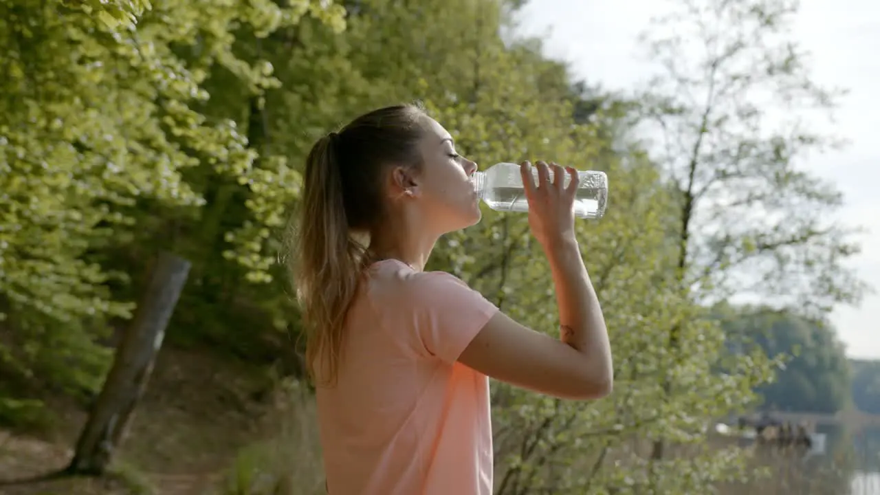 Woman drinking water from bottle standing by lake and woods