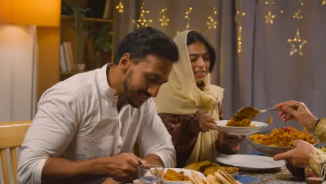 Muslim Family Sitting Around Table At Home With Woman Serving Biryani At Meal To Celebrate Eid