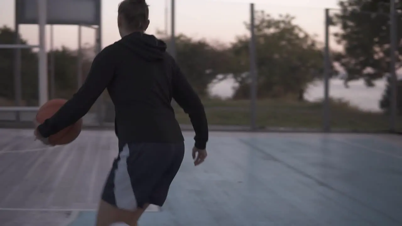 Backside Footage Of A Young Basketball Female Player Jogging With Ball On The Outdoors Court And Throw It To The Hoop