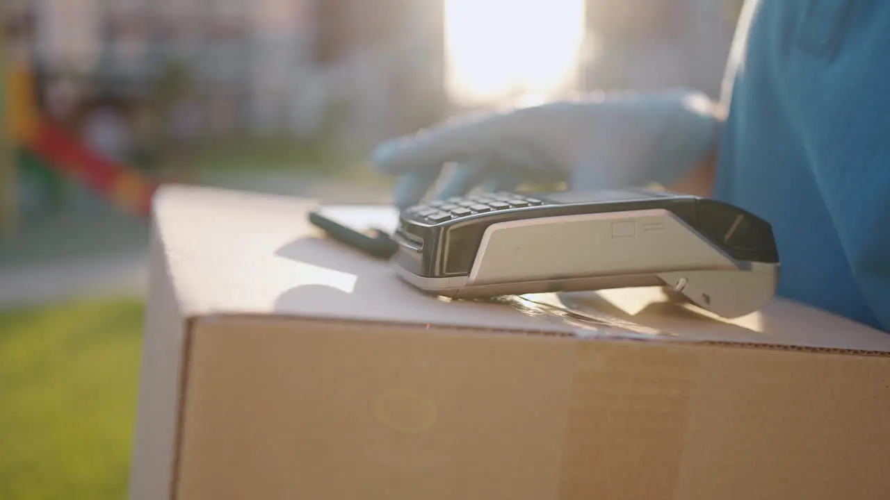 A male deliveryman in gloves brings a box and gives the terminal to pay for the purchase Carry a box with an NFC credit card payment terminal or mobile phone Contactless payment