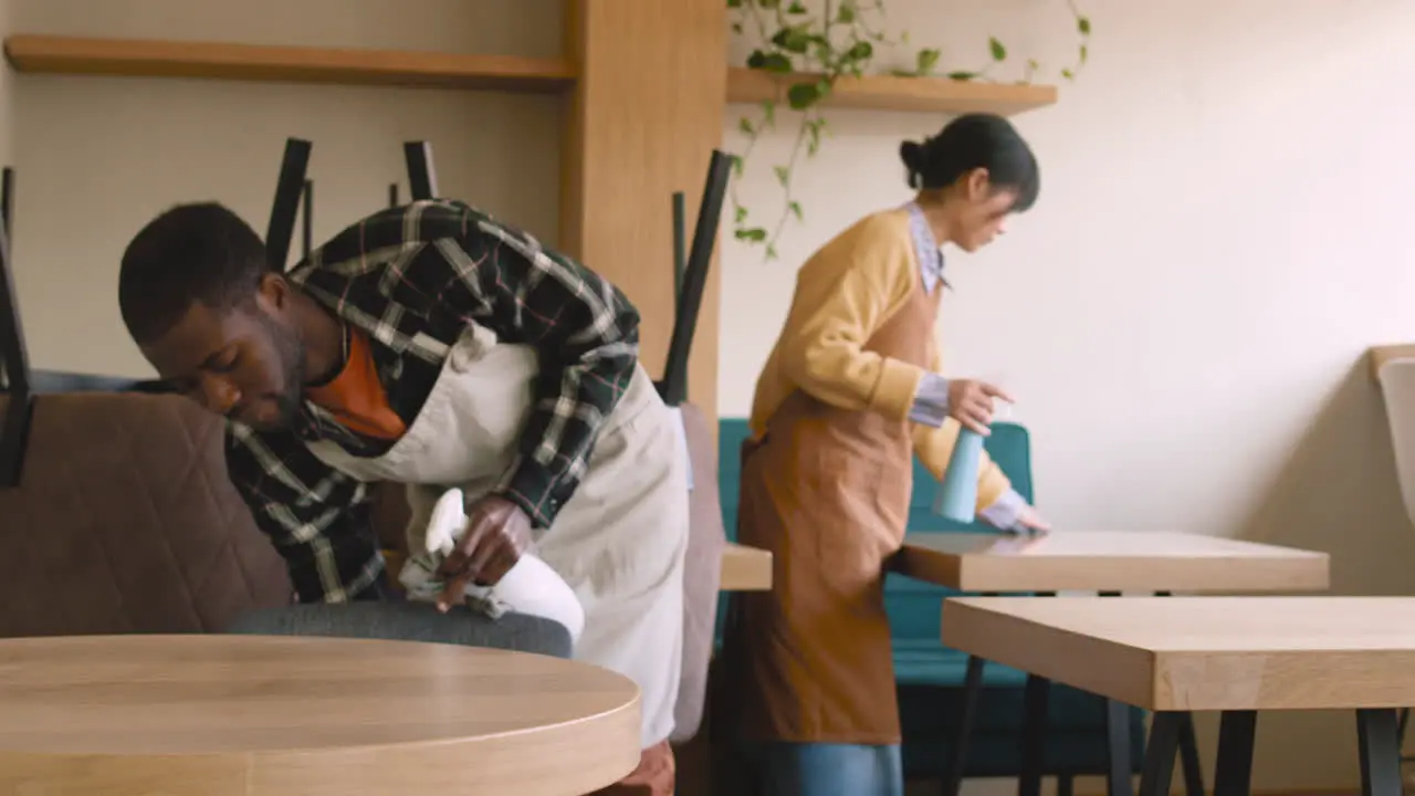 Waitress Cleaning Coffee Shop Table With Disinfectant Spray And Rag While Waiter Raising Chair To Arrange In Preparation For Closing Coffee Shop