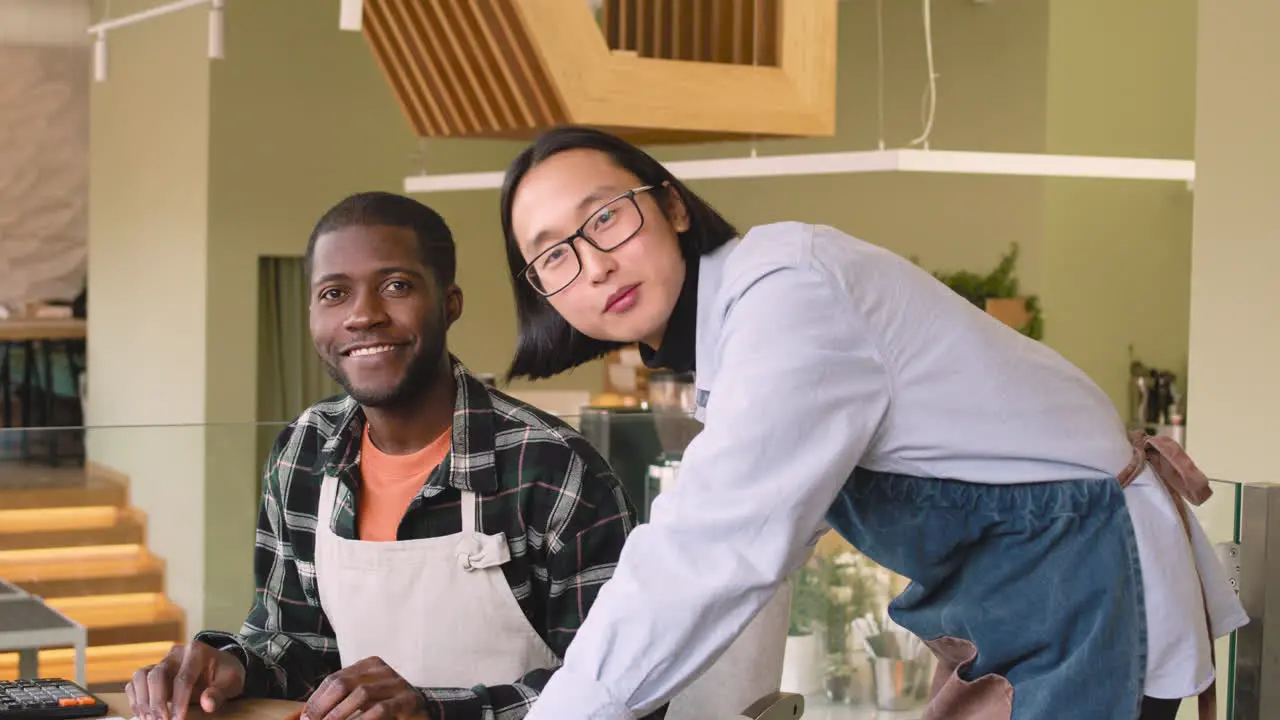 Two Multiethnic Coffee Shop Owners Smiling At Camera While Calculating Finance Bills On Laptop Computer