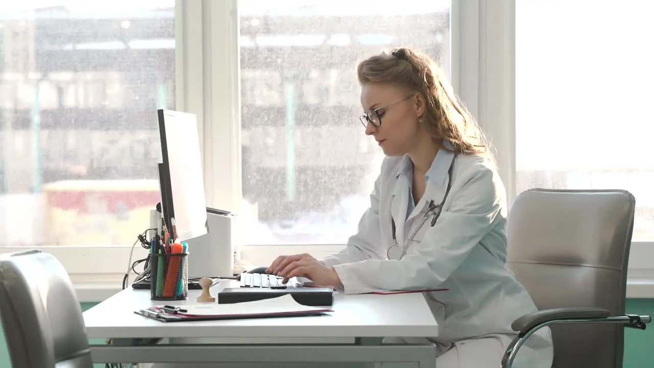 Female Doctor Working On Computer While Sitting At Desk In Her Consulting Room 1