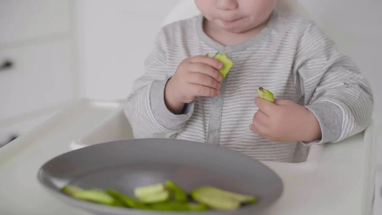 Close Up Of A Cute Baby Boy Eating Avocado Slices Sitting In High Chair In The Kitchen