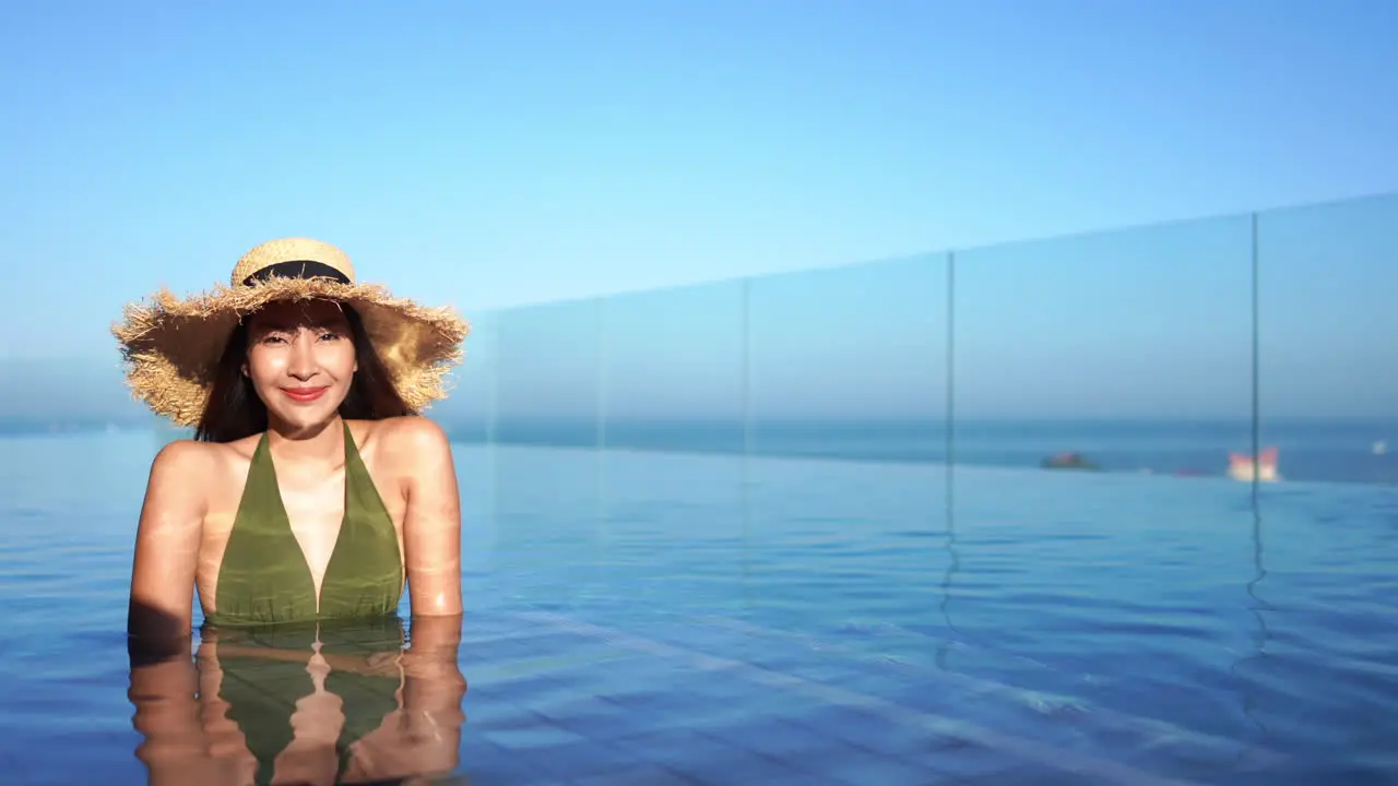 Pretty fit Asian woman in a green bathing suit and floppy straw sun hat relaxes in a resort luxury pool