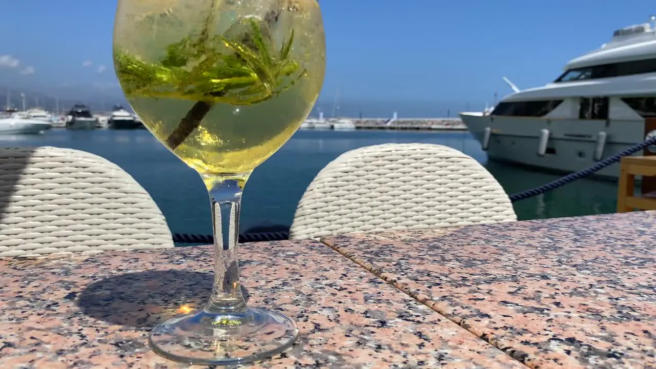 Fancy cocktail with champagne and rosemary by the sea in Puerto Banus with yachts luxury holiday destination drink on a tropical vacation with sea view in Marbella Spain 4K tilting up