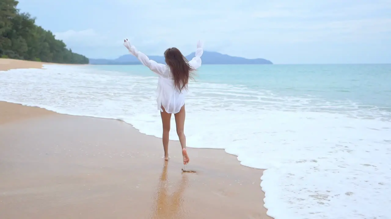 With her back to the camera a healthy fit woman wearing a bathing suit and a white shirt coverup runs along the surf as it rushes to the shore