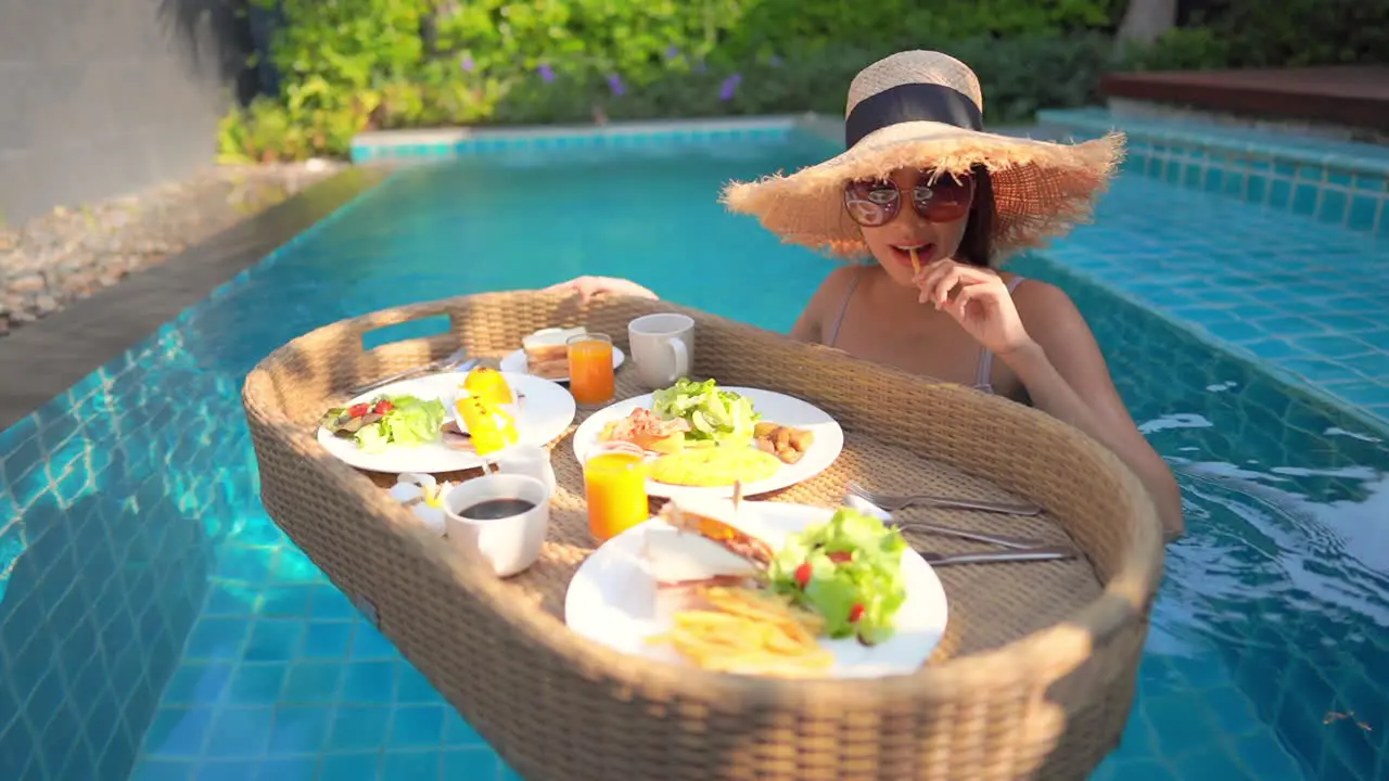 Asian woman takes a bite from her breakfast on a floating tray while in a swimming pool