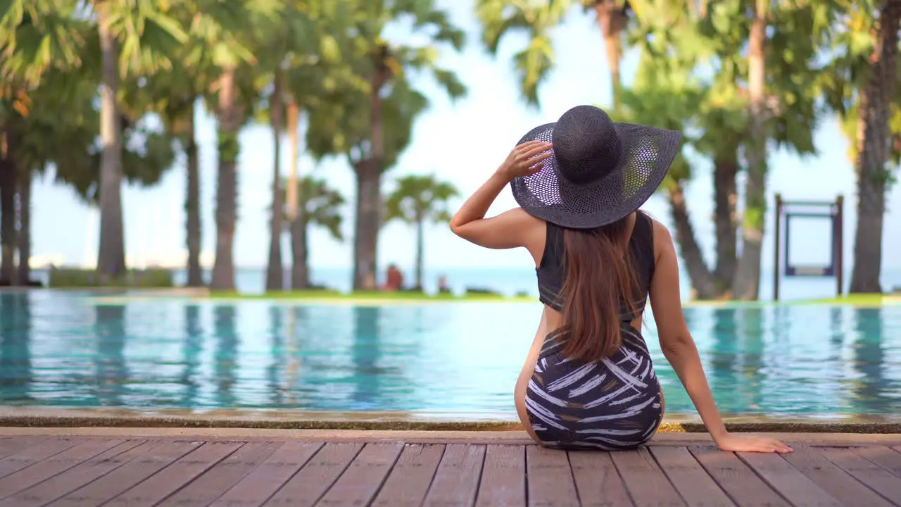 Beautiful long haired woman sits at the edge of pool wearing swimsuit and hat