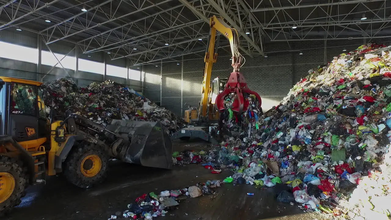 Digger and crane loading heap of trash in recycling yard hall aerial view