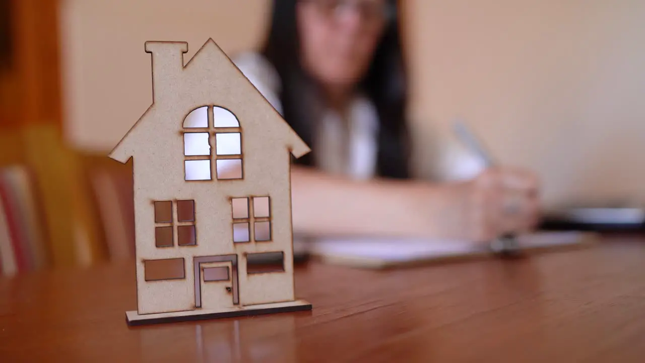 Professional Woman Finishing Paperwork in Blurry Background With Focus On Miniature House Icon On Office Table