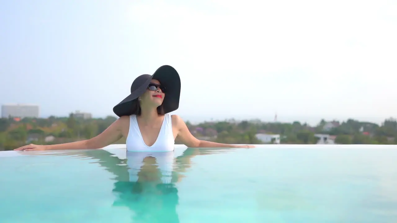 A woman in a huge black floppy sun hat leans along the edge of a swimming pool as she takes in the view