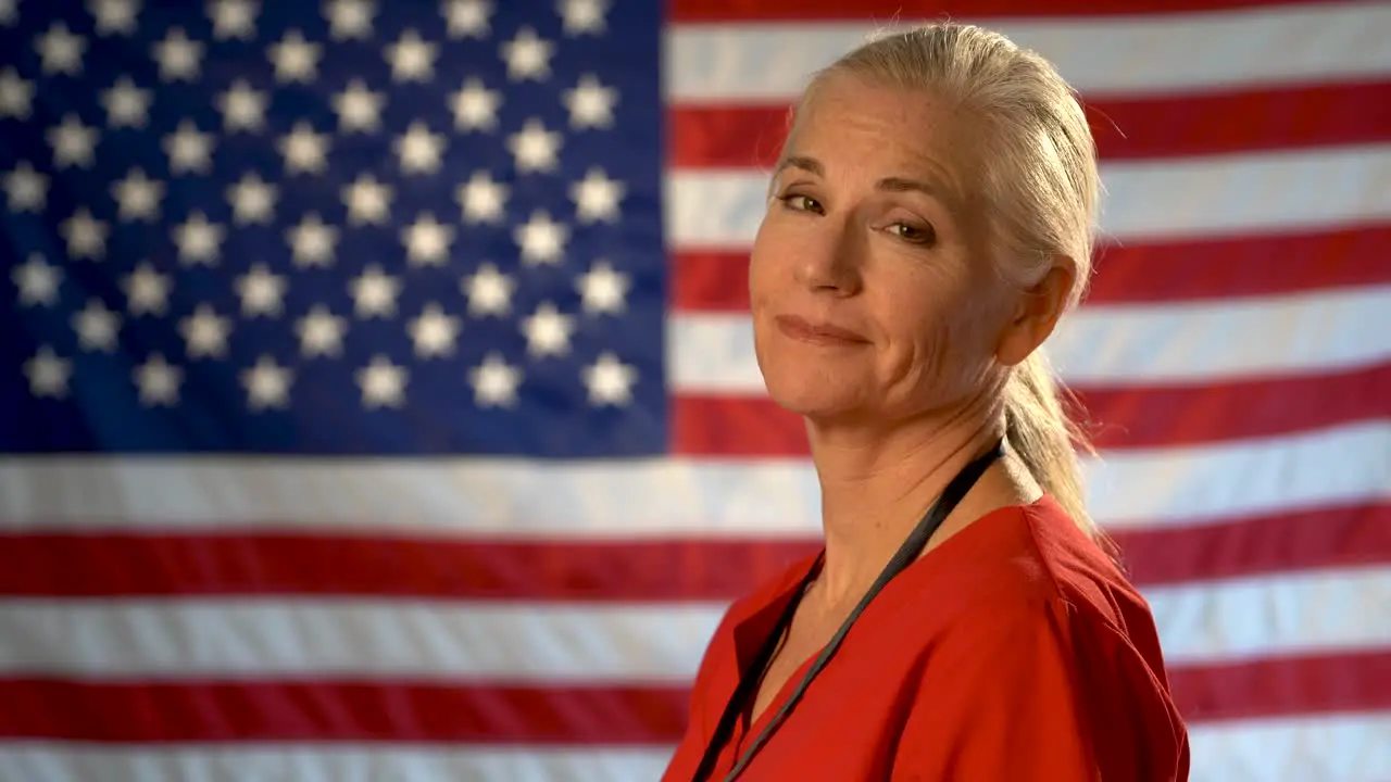 Medium tight portrait of the back of blonde nurses head as she turns and looks at camera looking relieved and happy with out of focus US flag