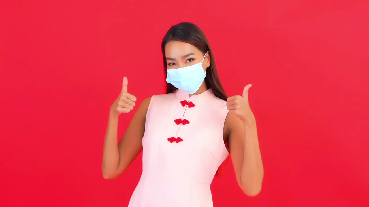 A young woman showing her face mask and gives thumbs up hands gesture covid-19 virus pandemic concept red background static shot