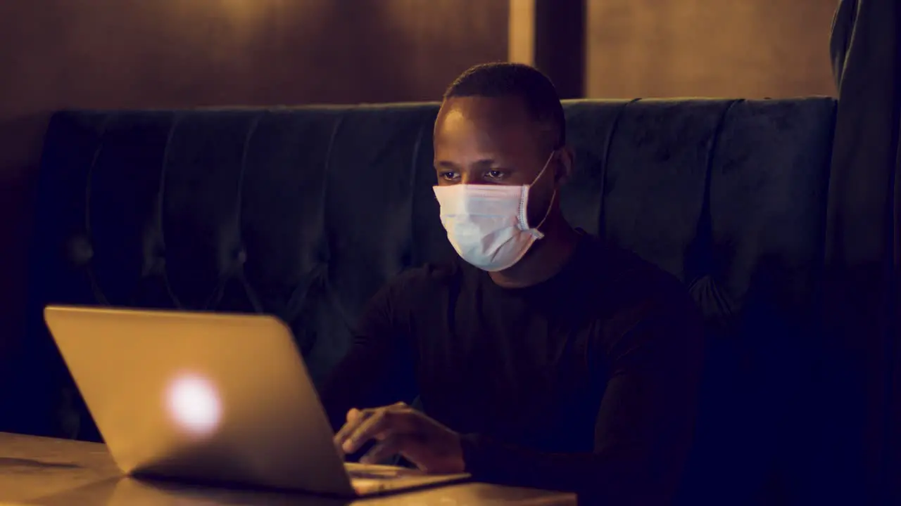 Male working on a laptop in a dark café wearing a facemask