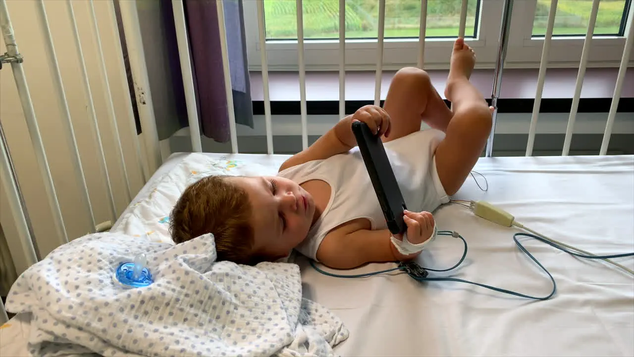 Young baby boy laying down in hospital bed watching movies on smartphone