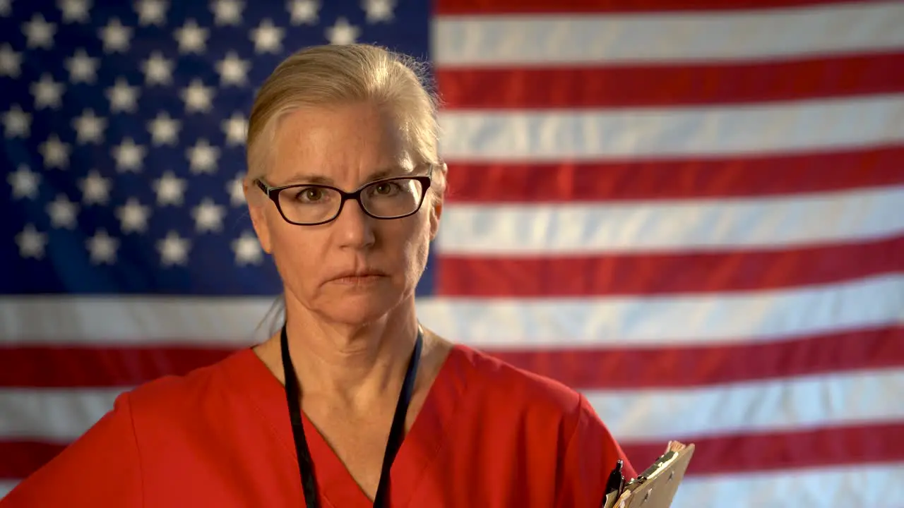 Medium tight portrait of a healthcare nurse with clipboard looking concerned and sad with an out of focus US flag