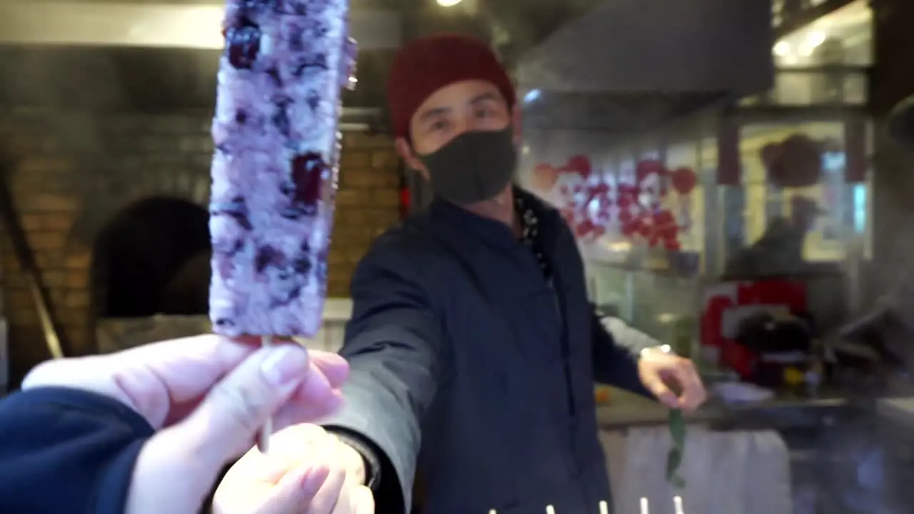 Person selling street food handing to the camera and a hand grabbing sticky rice and red beans on a stick