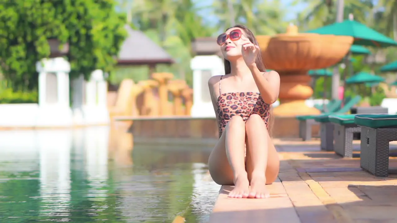 American Woman in Spotty Swimsuit Tanning Sitting on the Edge of the Swimming Pool in Exotic Tropical Hotel in Hawaii