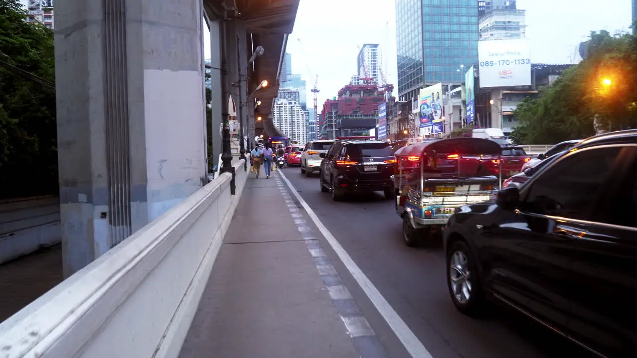 A man walking under a sky train taking videos of a particular street in Bangkok revealing cars motorcycles tuk-tuks busses buildings taxis and people walking