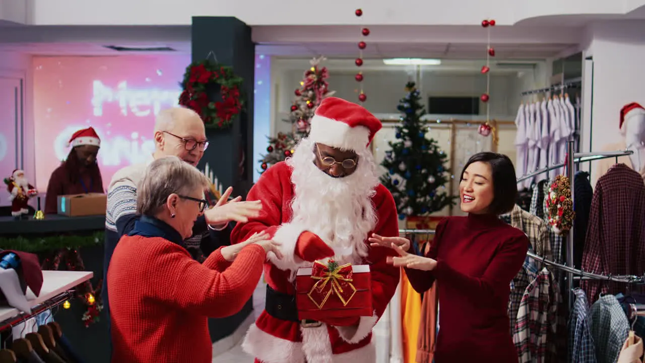 Retail assistant wearing Santa Claus suit in holiday adorn fashion boutique holding present box acting as raffle tickets container drawing random senior woman among happy clients