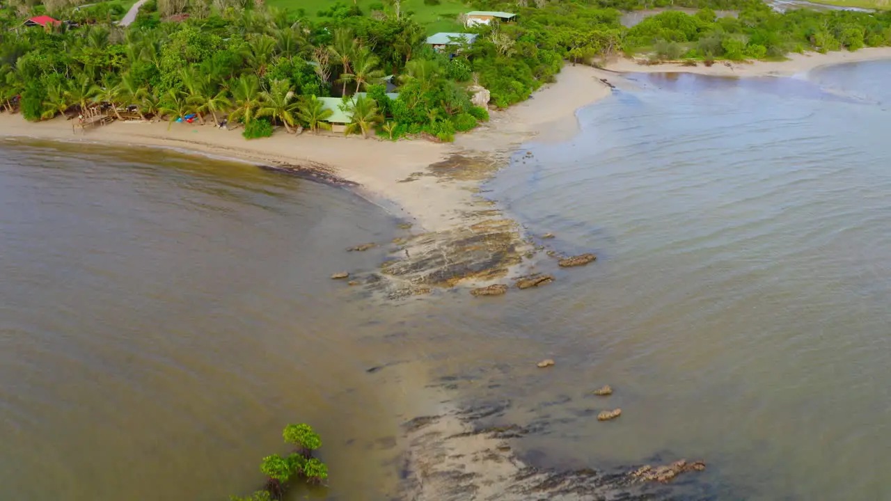 fly over a beach with a rock on the middle of the water toward a tropical wood with some traditional habitations scattered around