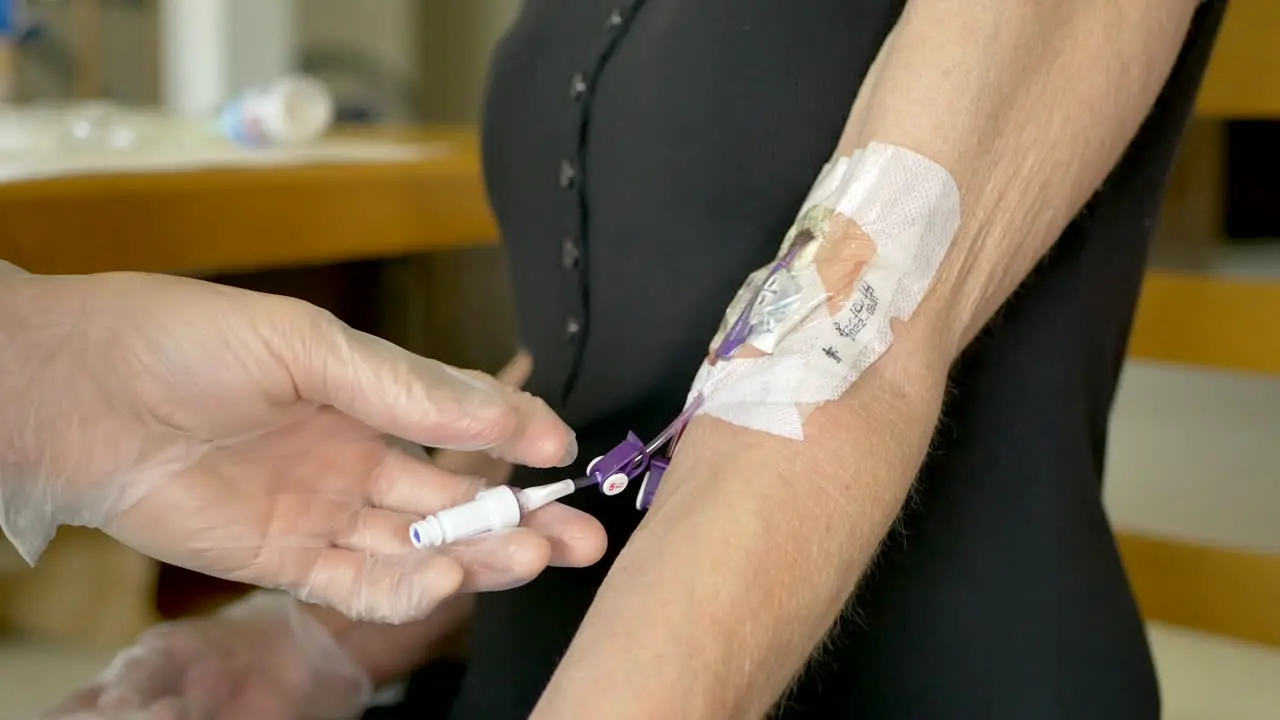 Medical nurse doing an at home iv patient visit wearing sterile gloves and injecting saline solution from a syringe into a picc line