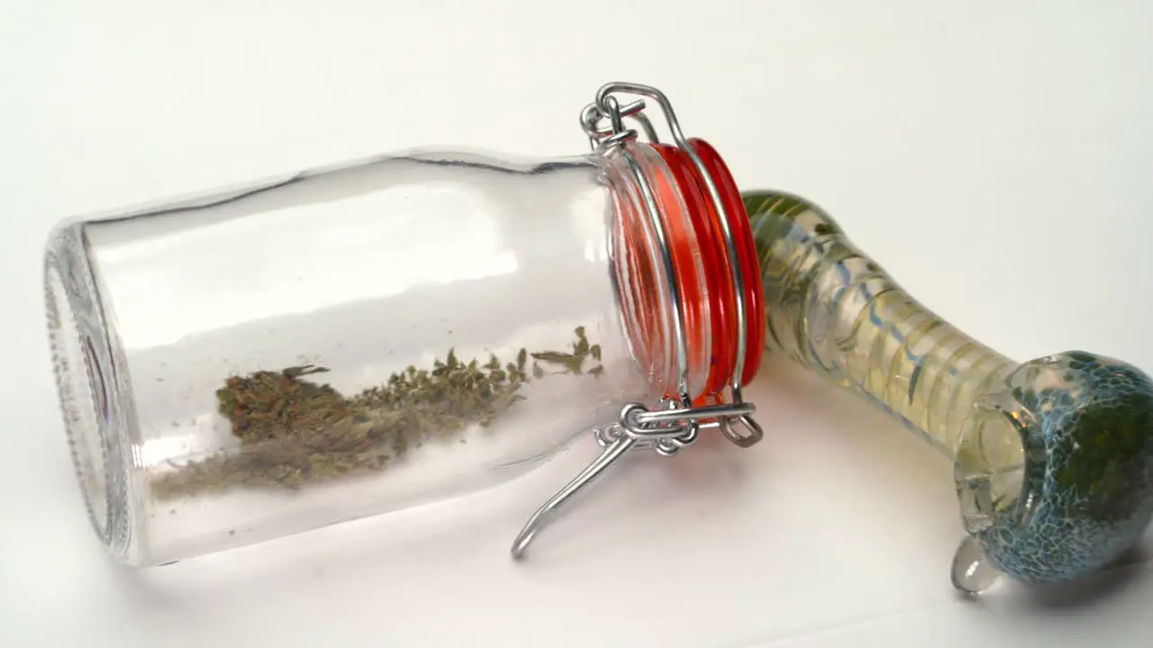 Pan across a clear bottle with marijuana grounds inside and glass smoking pipe